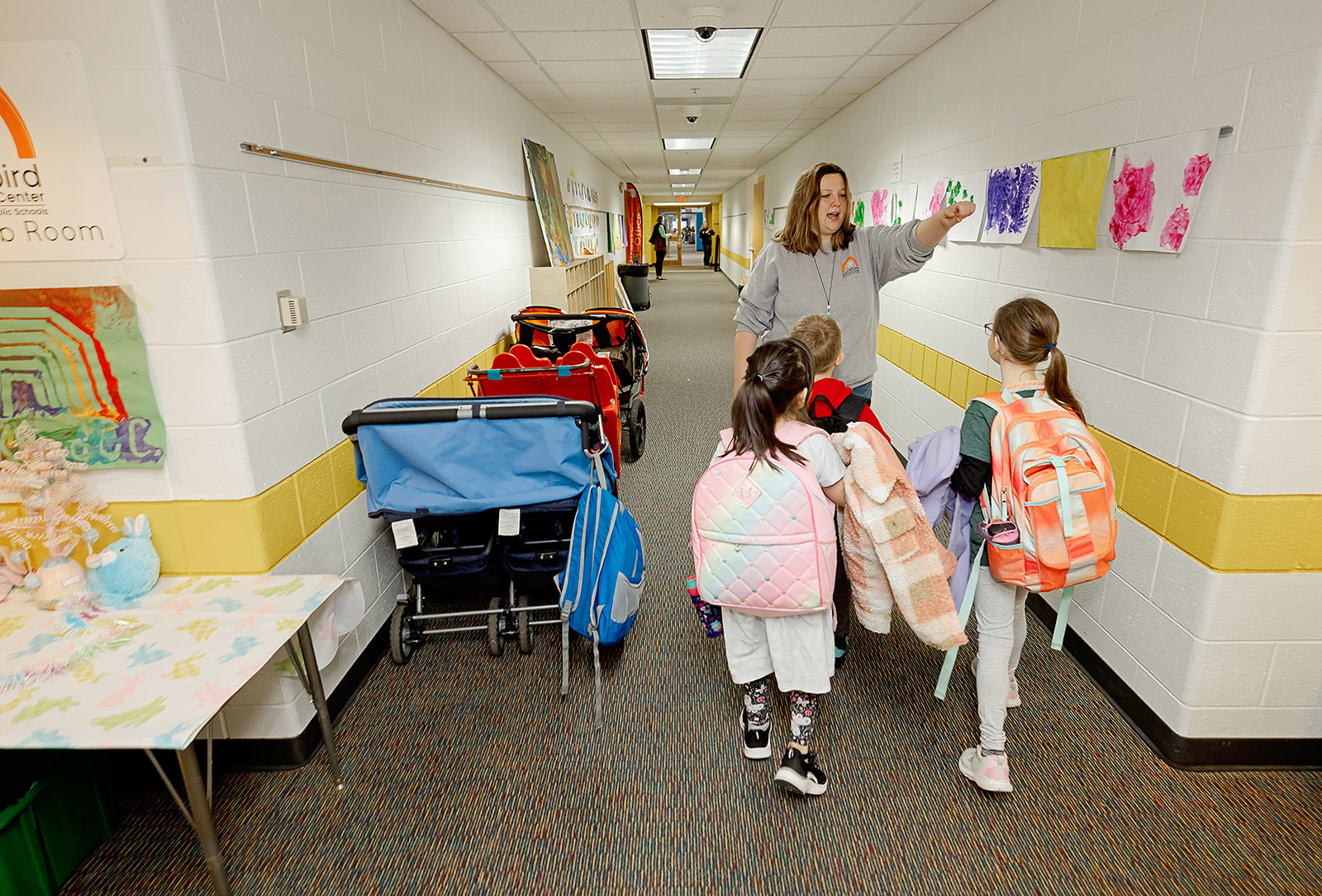 Staff member instructing three children carrying backpacks in a hallway.