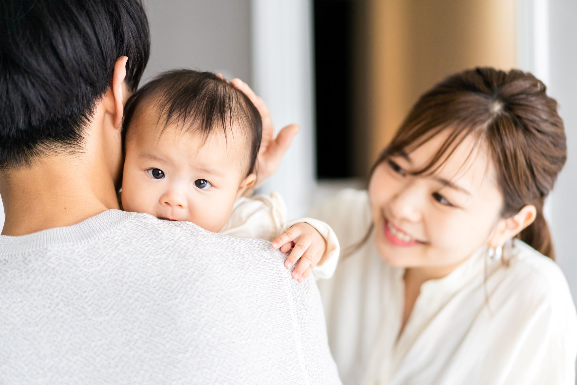 Family photo of one parent holding a baby while another parent smiles and touches the baby’s head.
