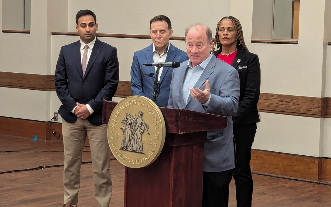 Detroit Mayor Mike Duggan speaks at a podium. Jeff Morenoff and others stand behind him.