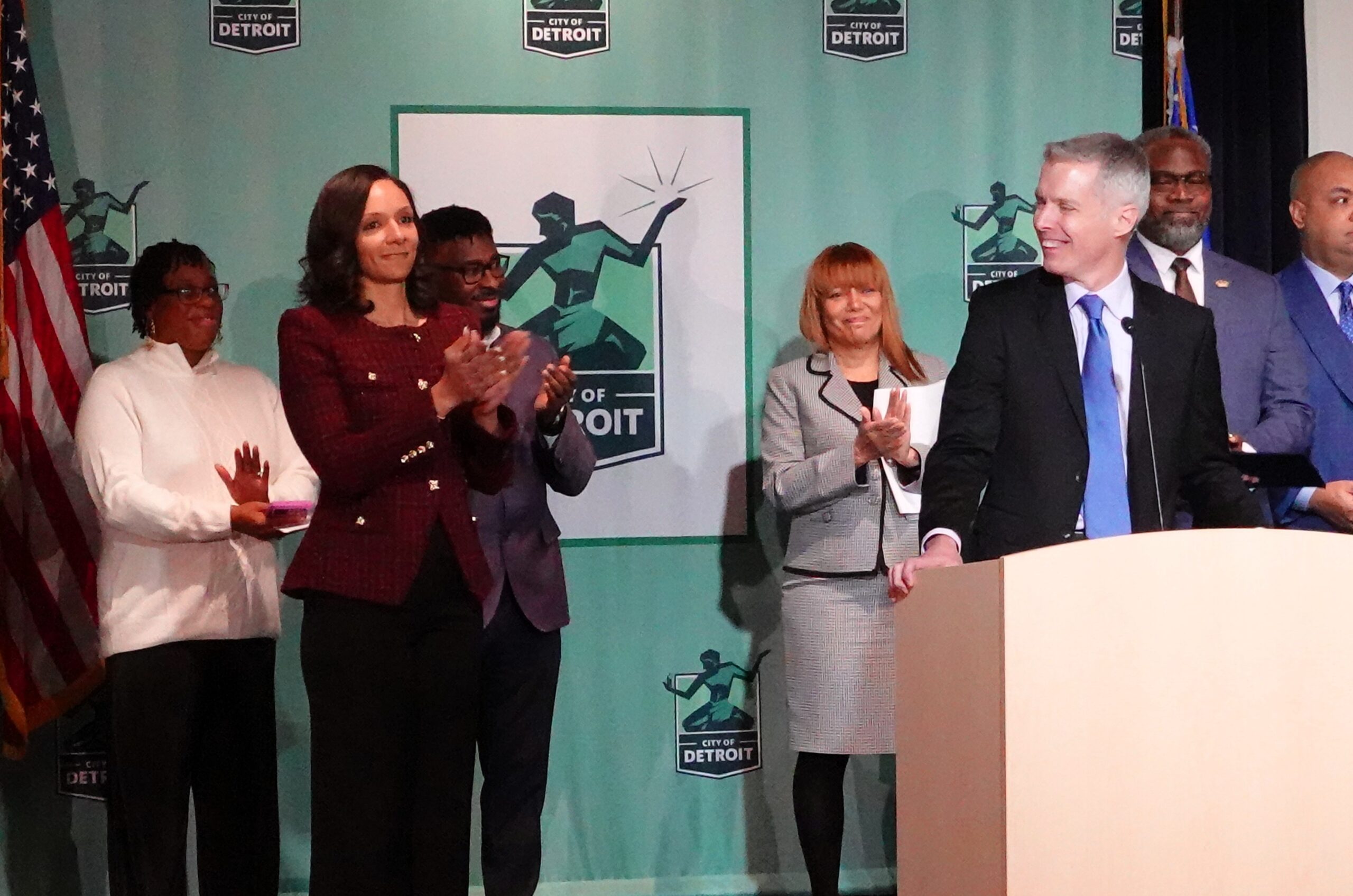 Graphic of Luke Shaefer speaks at a podium while Detroit Mayor Mary Sheffield stands nearby applauding